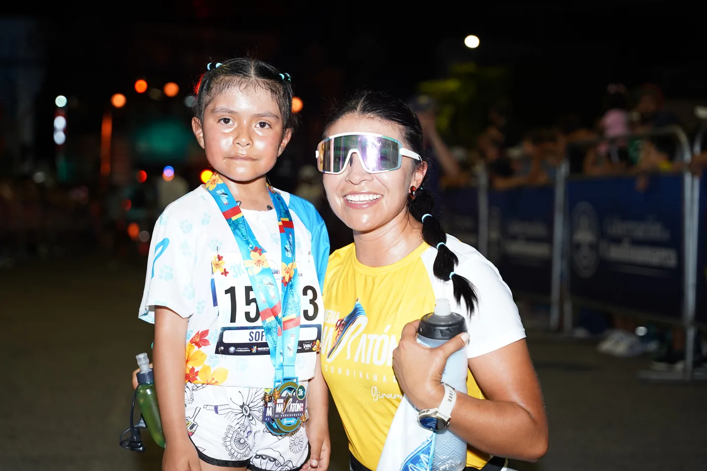Una niña con su medalla de finalista junto a una corredora, de noche, en la meta